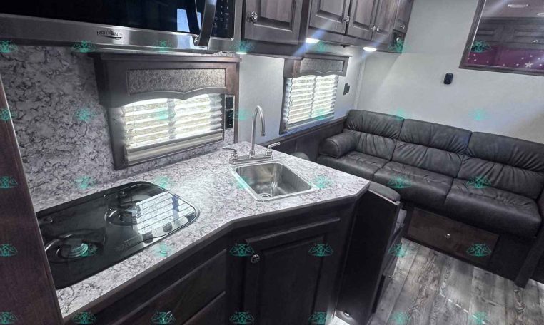 Interior of a modern RV kitchen with a marble-like countertop, stainless microwave, and dark wood cabinets above a sink and stove, next to a black leather sofa on the right (bright overhead lights).