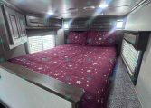 Interior of a travel trailer bedroom: a full/double bed with purple star-pattern bedding, dark wood overhead cabinets, and two small windows on each side.