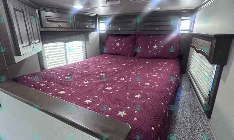Interior of a travel trailer bedroom: a full/double bed with purple star-pattern bedding, dark wood overhead cabinets, and two small windows on each side.