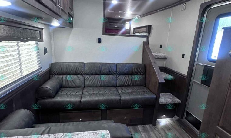 Interior of a compact RV living area with a dark leather sofa, overhead cabinets, and a kitchen counter to the right; bright ceiling lights and vents above.