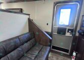 Interior of a trailer living area with a dark leather sofa along a partition wall and a doorway with a windowed metal frame on the right.