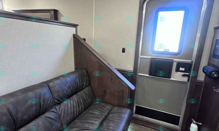 Interior of a trailer living area with a dark leather sofa along a partition wall and a doorway with a windowed metal frame on the right.