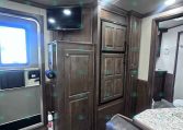 Interior of a dark wood RV kitchen with a mounted flat-screen TV above a cabinet and a tall refrigerator next to it