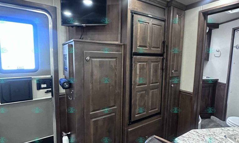 Interior of a dark wood RV kitchen with a mounted flat-screen TV above a cabinet and a tall refrigerator next to it