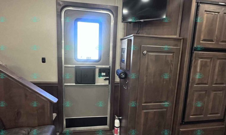 Interior of a wood-loaded trailer living area with a mounted TV, entry door, tall cabinets, and a fire extinguisher nearby.