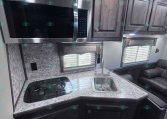 Compact RV kitchen with stainless microwave above a granite-like counter, two-burner stove, and sink under dark wood cabinets
