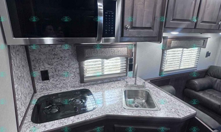 Compact RV kitchen with stainless microwave above a granite-like counter, two-burner stove, and sink under dark wood cabinets