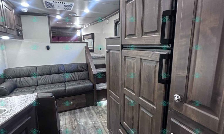 Interior of a modern RV with dark wood cabinets, a gray leather sofa, and a tall double-door refrigerator, view from the kitchen area.