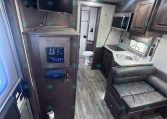 Interior of an RV with a mounted TV above a cabinet, followed by a kitchenette: microwave, sink, stove, and dark wood cabinets; a black leather sofa nearby.