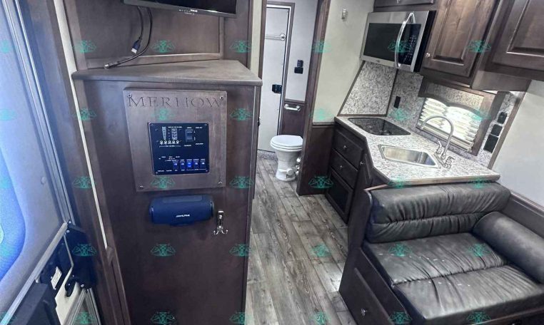 Interior of an RV with a mounted TV above a cabinet, followed by a kitchenette: microwave, sink, stove, and dark wood cabinets; a black leather sofa nearby.
