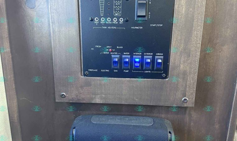 Metal outer panel with the word 'MERHOW' carved or engraved at the top, featuring a black control board with switches and indicators beneath it (likely a trailer or RV control panel). Inset readings show tank/level indicators and a toggle switch labeled 'START/STOP' nearby, plus a blue Alpine speaker mounted below.