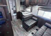 Interior of a modern RV/travel trailer: kitchen with countertop, double sink, microwave, and dark wood cabinets, next to a leather sofa area