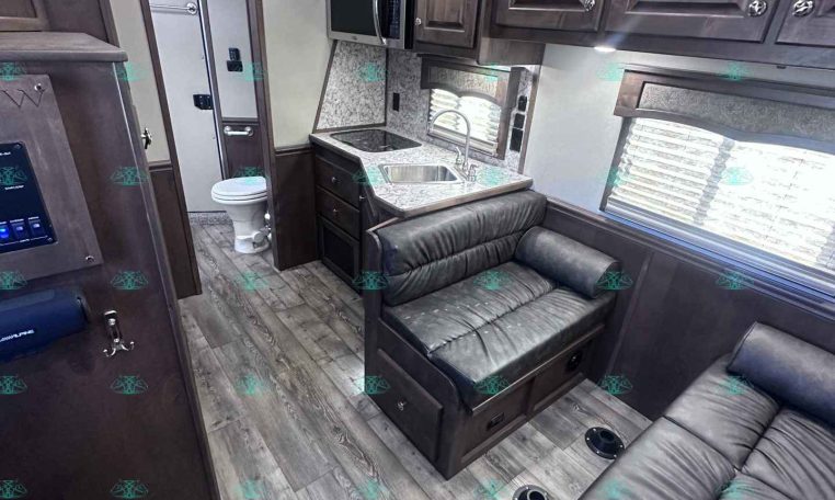 Interior of a modern RV/travel trailer: kitchen with countertop, double sink, microwave, and dark wood cabinets, next to a leather sofa area