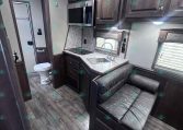 Interior of a modern RV kitchen with microwave, dark wood cabinets, granite countertop, sink, and lounge seating nearby; bathroom door visible in background.