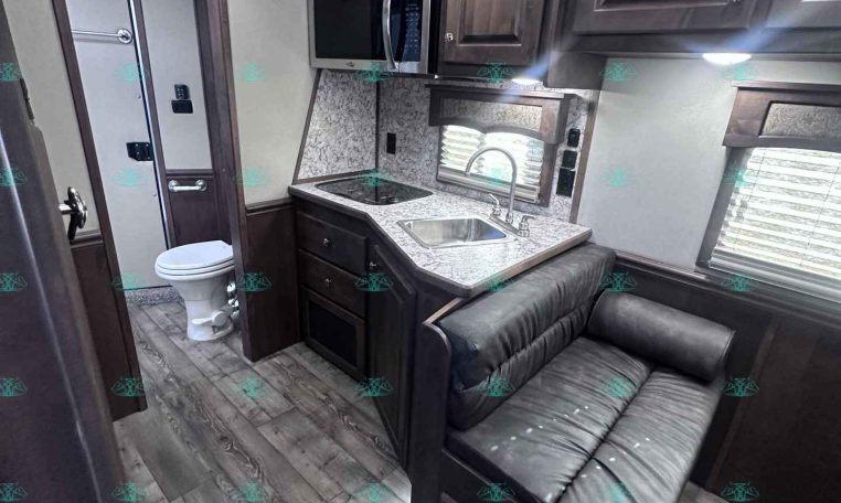 Interior of a modern RV kitchen with microwave, dark wood cabinets, granite countertop, sink, and lounge seating nearby; bathroom door visible in background.