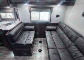 RV living area with dark wood overhead cabinets, a gray L/U-shaped sofa, and a window with blinds above a compact kitchen sink on the left.