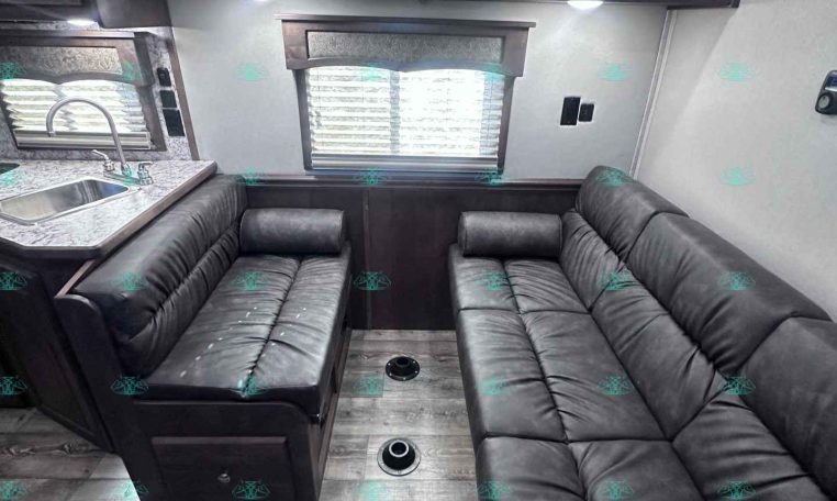 RV living area with dark wood overhead cabinets, a gray L/U-shaped sofa, and a window with blinds above a compact kitchen sink on the left.