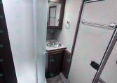 Compact RV bathroom with a curved glass shower on the left, small vanity with sink and mirror cabinet on the back wall, and a door on the right.