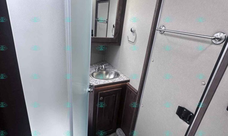 Compact RV bathroom with a curved glass shower on the left, small vanity with sink and mirror cabinet on the back wall, and a door on the right.