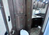 RV bathroom and kitchenette area with dark wood cabinets, a toilet in the foreground, and a sink with counter visible in the back.