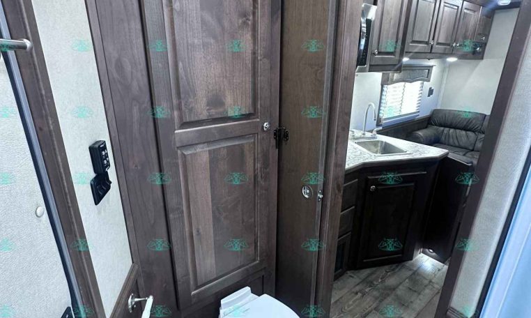 RV bathroom and kitchenette area with dark wood cabinets, a toilet in the foreground, and a sink with counter visible in the back.