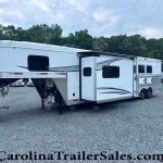 White fifth-wheel RV with an extended slide-out parked on a gravel lot, with a tree line in the background.