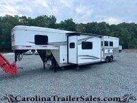 White fifth-wheel RV with an extended slide-out parked on a gravel lot, with a tree line in the background.