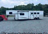 White horse trailer with windows parked on a gravel lot, with a red tractor attachment on the left and green trees in the background.