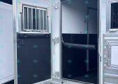 Open aluminum trailer with both doors ajar, showing a small barred window on the left door and a ramped rear entry onto a gravel surface.