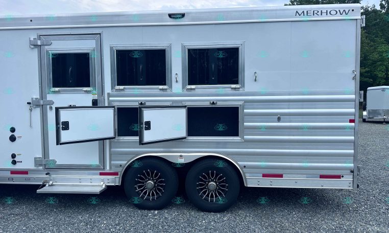 Silver Merhow horse trailer with two small side windows, a side entry door open, and fold-out lower steps on a gravel lot with tandem wheels.