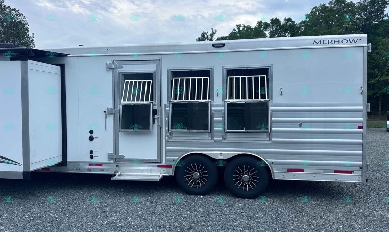 Side view of a silver Merhow horse trailer with barred windows, two axles, and a gravel lot in front of a treeline and cloudy sky.