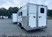 Rear and side view of a large silver Merhow horse trailer on a gravel lot, with trees in the background and another trailer visible in the distance.