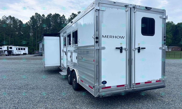 Rear and side view of a large silver Merhow horse trailer on a gravel lot, with trees in the background and another trailer visible in the distance.