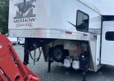 Silver horse-trailer with Merhow Stampede logo, side window, and exposed propane tanks beneath; red hydraulic machinery nearby in a gravel lot.