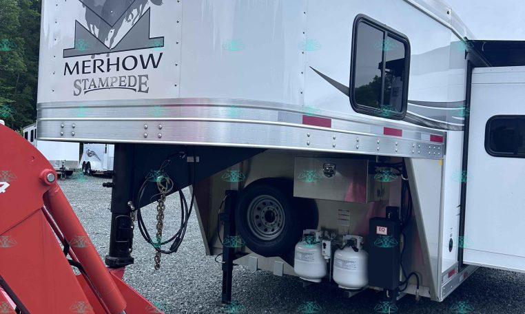 Silver horse-trailer with Merhow Stampede logo, side window, and exposed propane tanks beneath; red hydraulic machinery nearby in a gravel lot.