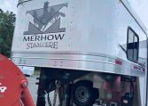 Rear side of a silver Merhow Stampede horse trailer with a stylized emblem of horses and mountains and a curved top edge in a parking area.