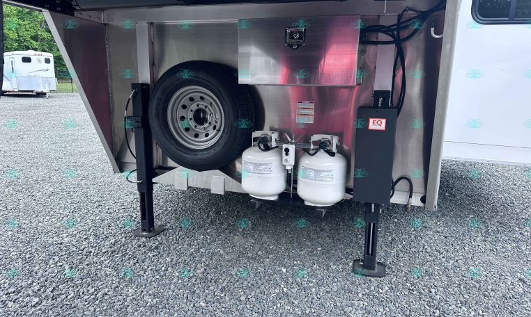 Open trailer storage area with a mounted spare tire, two propane tanks, hoses, and a metal storage box visible inside the compartment.
