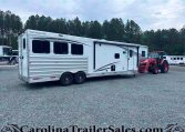 White Merhow horse trailer with two large side windows, parked on a gravel lot beside a red tractor, with pine trees and a cloudy sky in the background.