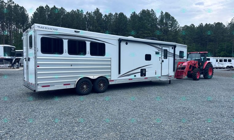 White Merhow horse trailer with two large side windows, parked on a gravel lot beside a red tractor, with pine trees and a cloudy sky in the background.