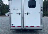 Rear view of a silver Merhow trailer with double doors, a small rounded window, and metal latches on a gravel lot with trees in the background.