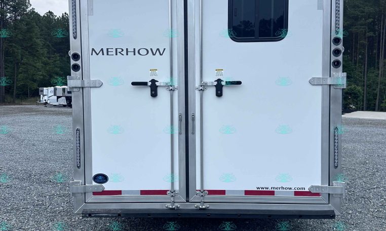 Rear view of a silver Merhow trailer with double doors, a small rounded window, and metal latches on a gravel lot with trees in the background.