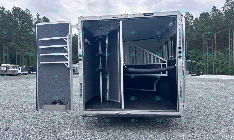 Open silver horse trailer with interior stalls and a ramp, parked on a gravel lot with trees in the background; watermark CarolinaTrailerSales.com at bottom.