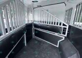 Interior of a white horse trailer with metal stall dividers and black padded lower walls, showing a narrow aisle and steps at the back.