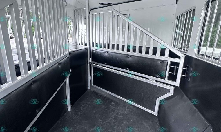 Interior of a white horse trailer with metal stall dividers and black padded lower walls, showing a narrow aisle and steps at the back.