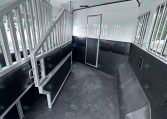 Interior of a white horse trailer with metal dividing stalls, padded lower walls, and a doorway seen from the aisle.