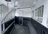 Interior of empty horse trailer with divider rails, gray walls, and barred windows on right; black rubber floor and ceiling lighting visible.