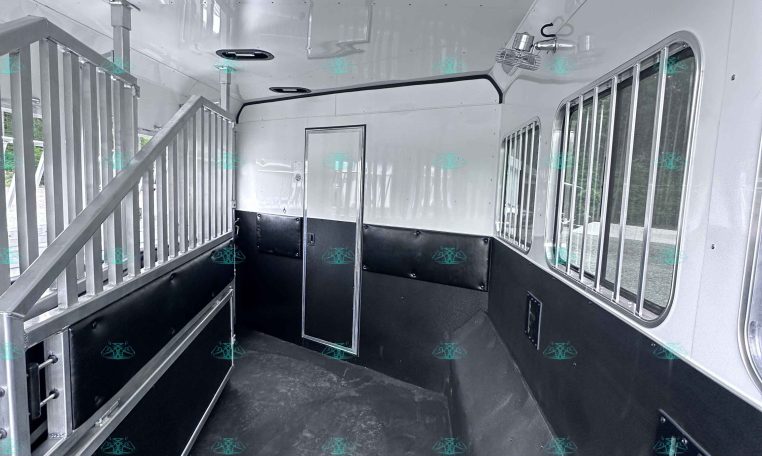 Interior of empty horse trailer with divider rails, gray walls, and barred windows on right; black rubber floor and ceiling lighting visible.