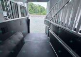 Interior of an aluminum horse trailer with a metal partition on the right, windows with bars along the left wall, and an open rear doorway revealing gravel and trees outside.