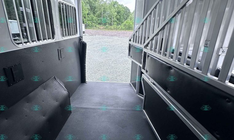 Interior of an aluminum horse trailer with a metal partition on the right, windows with bars along the left wall, and an open rear doorway revealing gravel and trees outside.