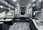 Interior view of a modern RV showing a kitchen with double sinks, cabinets, and a microwave on the right, and a seating area with a table on the left